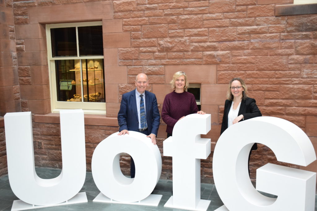 Professor David Clark, Dr Marian Krawczyk, and Dr Naomi Richards standing together in the foyer of Rutherford McCowan, next to the large U of G letters